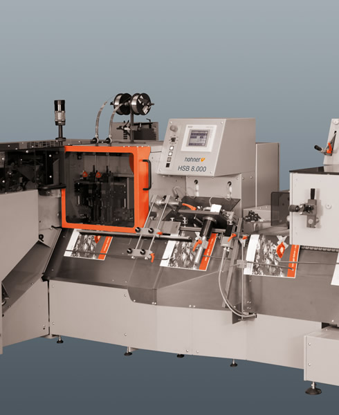 Official legacy studio render (490x600, dated 2012-04) of the HSB 8.000 tool-less saddle stitcher showing the stitching unit (orange knob), wire spools and inline feed table. ANCHORED machine (SC-HO-01).