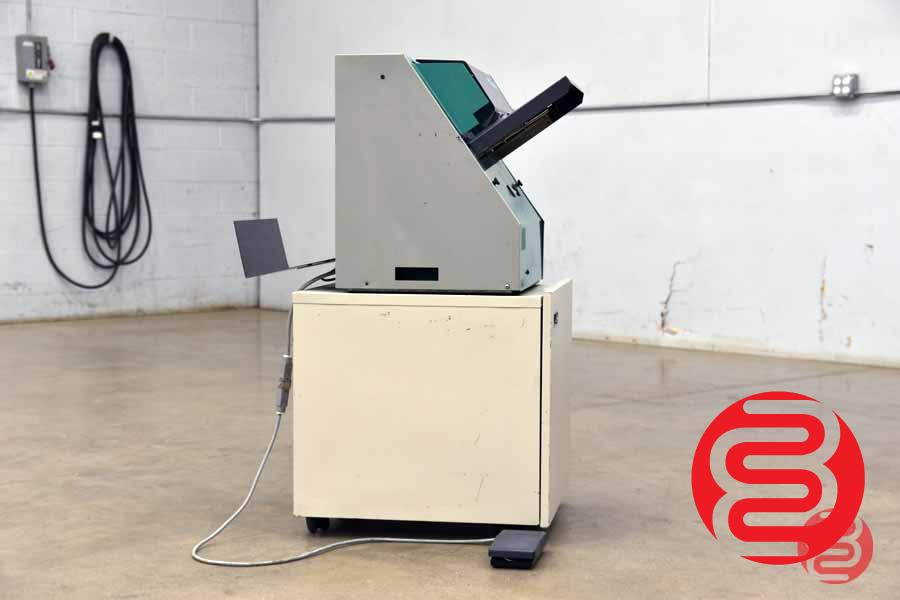 Side view of an early tabletop bookletmaker on a cabinet stand with foot pedal. HEAVY red circular dealer watermark; NO visible Nagel branding in frame. Weakest provenance — medium/low confidence it is the Foldnak-2. Flag for review / consider reject.