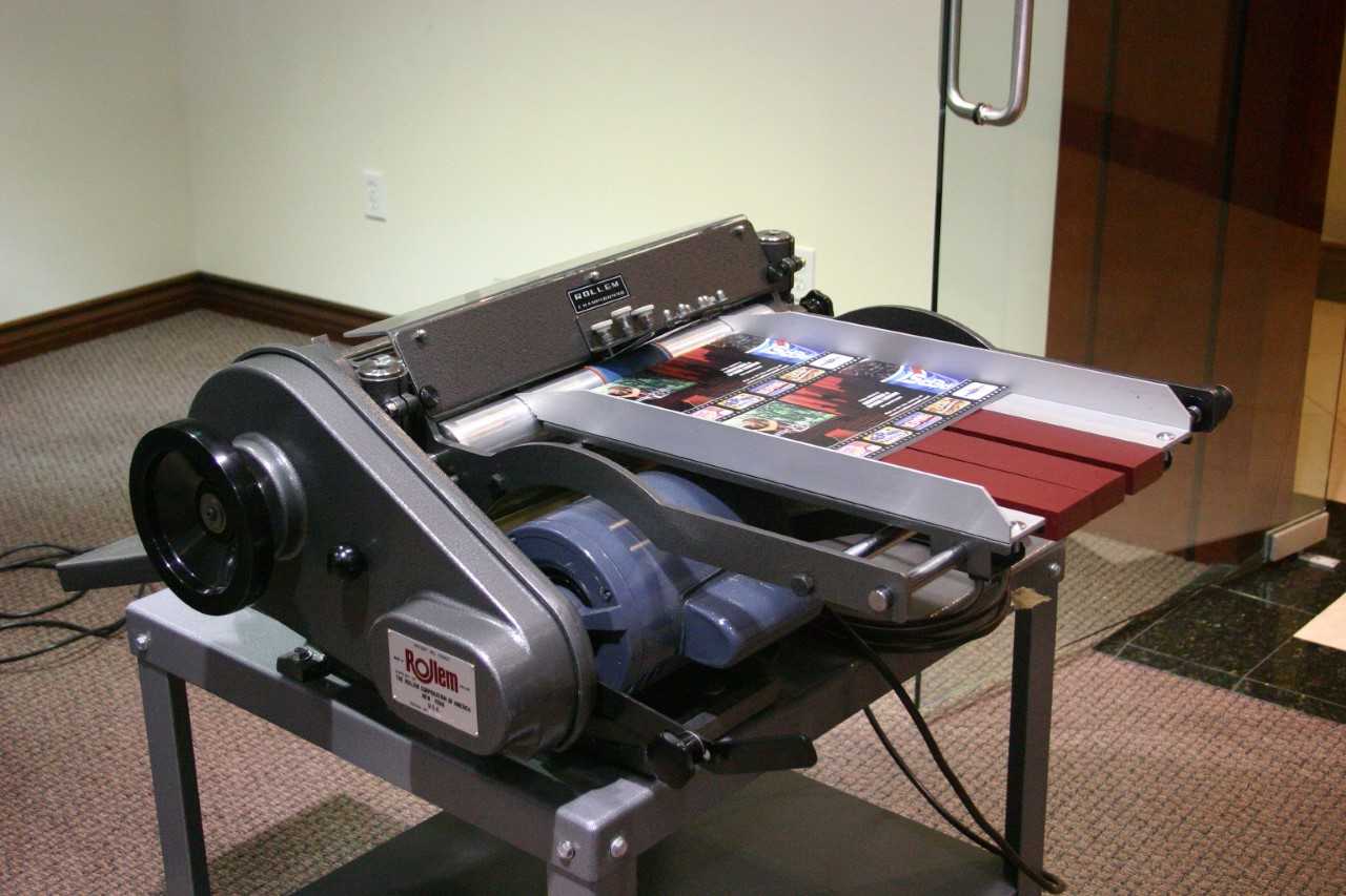 Official Rollem photo of the friction-fed Champion 990 SSP entry machine, printed sheet passing over the cross-shaft heads on the bench-top unit. Shows the shaft/head mounting deck clearly.